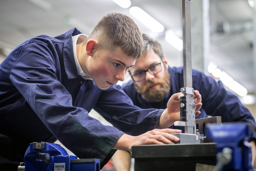 A lecturer and a student in navy-blue overalls working together in a mechanical engineering workshop. The lecturer is demonstrating how to use a tool on a metal component held in a vise.