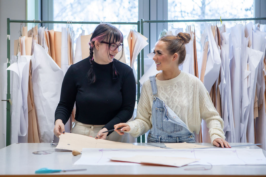 Two fashion students laughing while working on design patterns in a classroom.