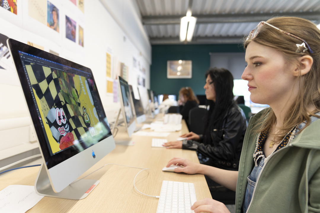 Student creating digital artwork on a desktop computer in a classroom with creative posters on the wall.