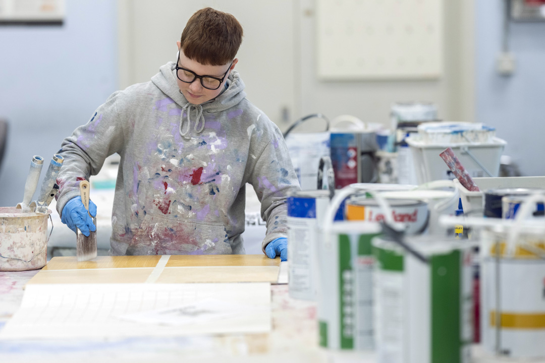 Focused student in paint-splattered hoodie painting on a wooden board