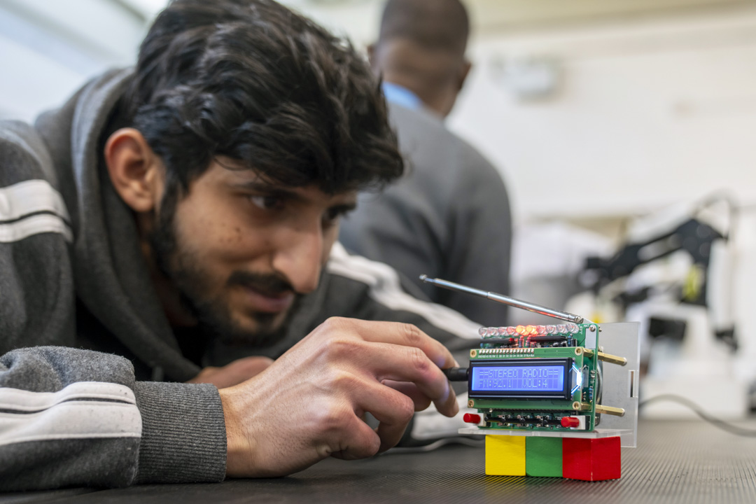 Student closely examining a custom-built radio circuit board with LED indicators and an antenna in an electronics lab