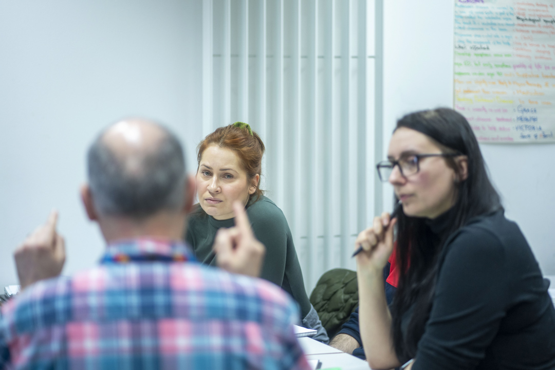 Lecturer leads a class discussion with students seated around a table and a population graph displayed behind.