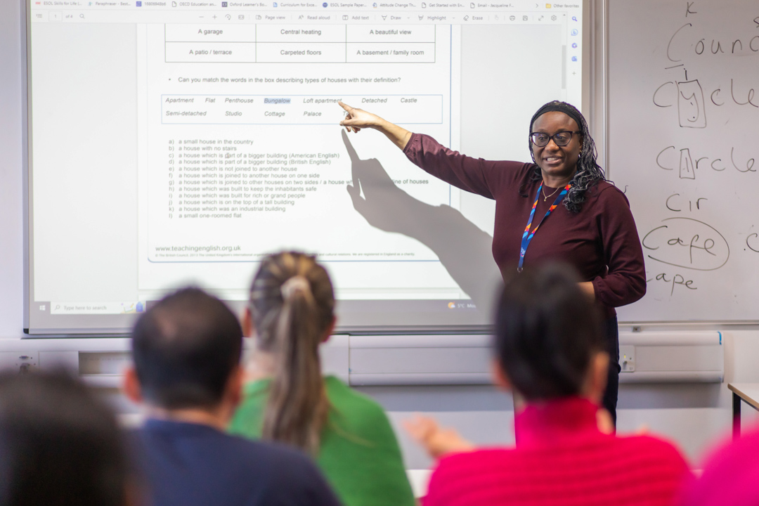 A female lecturer wearing glasses and a lanyard points to a projected lesson on a whiteboard while teaching a group of students.