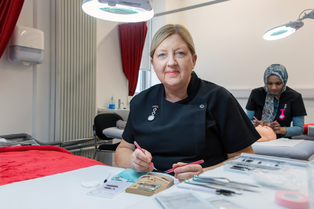 A beauty therapy student applies eyelash extensions to a mannequin head under a magnifying lamp in a professional training environment.