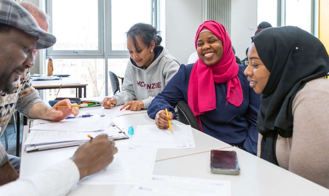 A diverse group of students, including a woman in a pink hijab, laughs and collaborates while working on an assignment in a classroom.