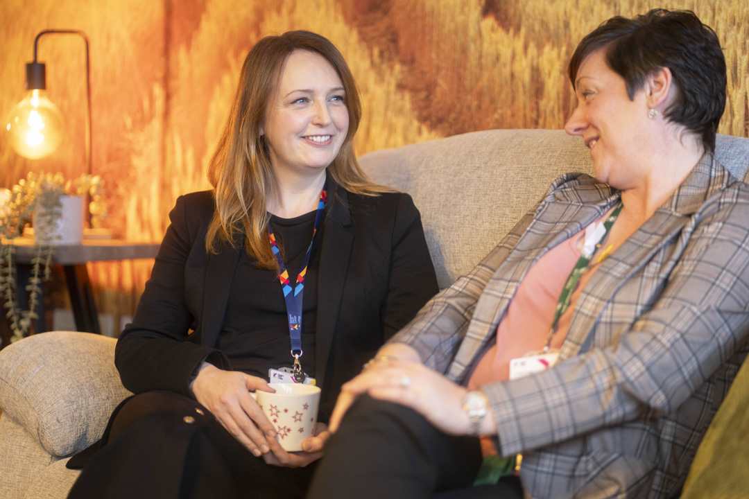 Two Glasgow Kelvin College staff members sitting on a sofa, smiling and chatting in a relaxed setting with warm lighting