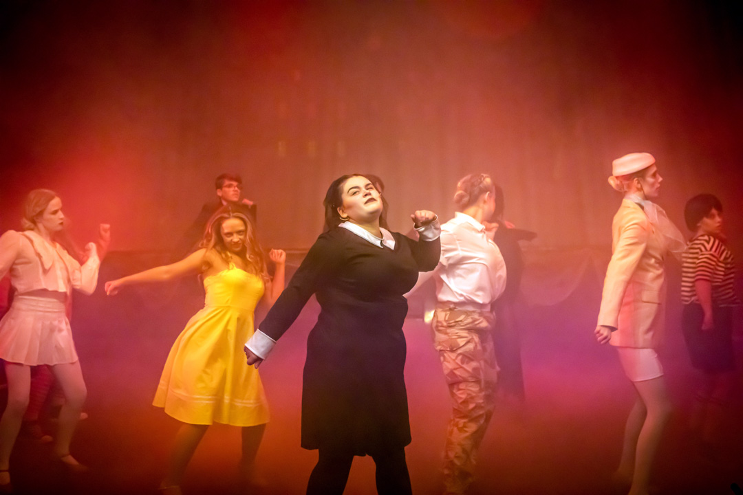 Musical theatre students dancing in formation during a stage performance, wearing contrasting costumes and performing under warm, atmospheric lighting.