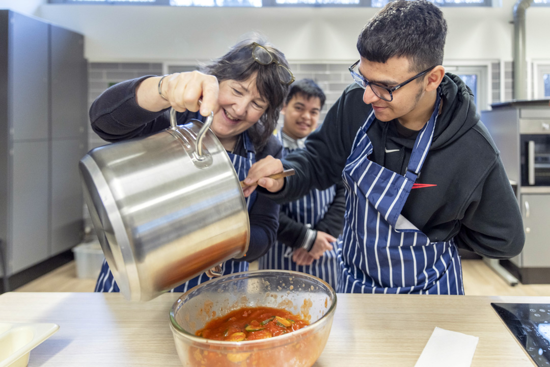 Woman pouring sauce from a large pot into a bowl while smiling and working with two young men in striped aprons.