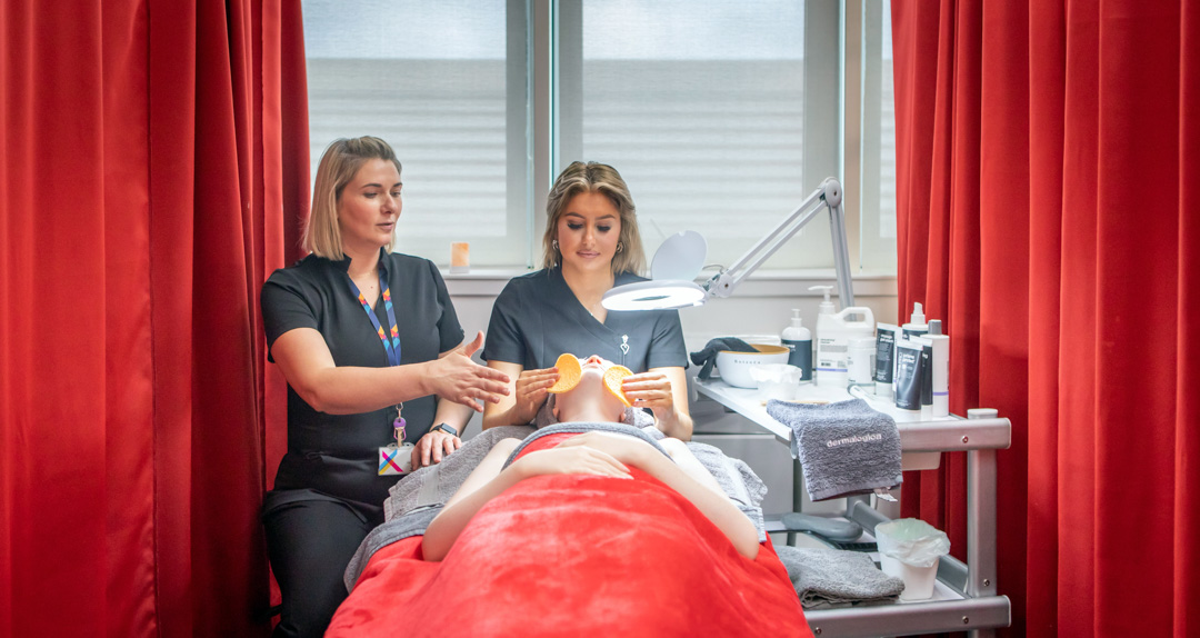 A lecturer demonstrates a facial massage technique to a beauty therapy student who is performing a treatment on a mannequin in a spa-like environment.