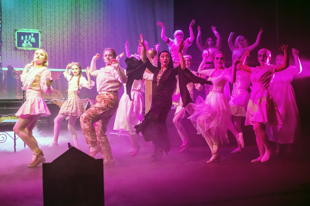 Musical theatre students performing a high-energy ensemble dance on stage, with raised arms, coordinated movement, and purple lighting.