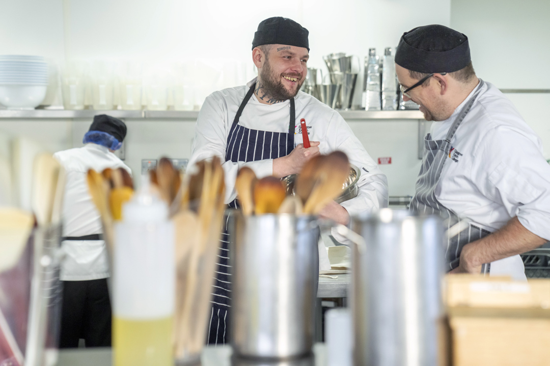 A chef with tattoos and a striped apron smiles while chatting with an instructor in a busy kitchen, with cooking utensils and supplies in the foreground.
