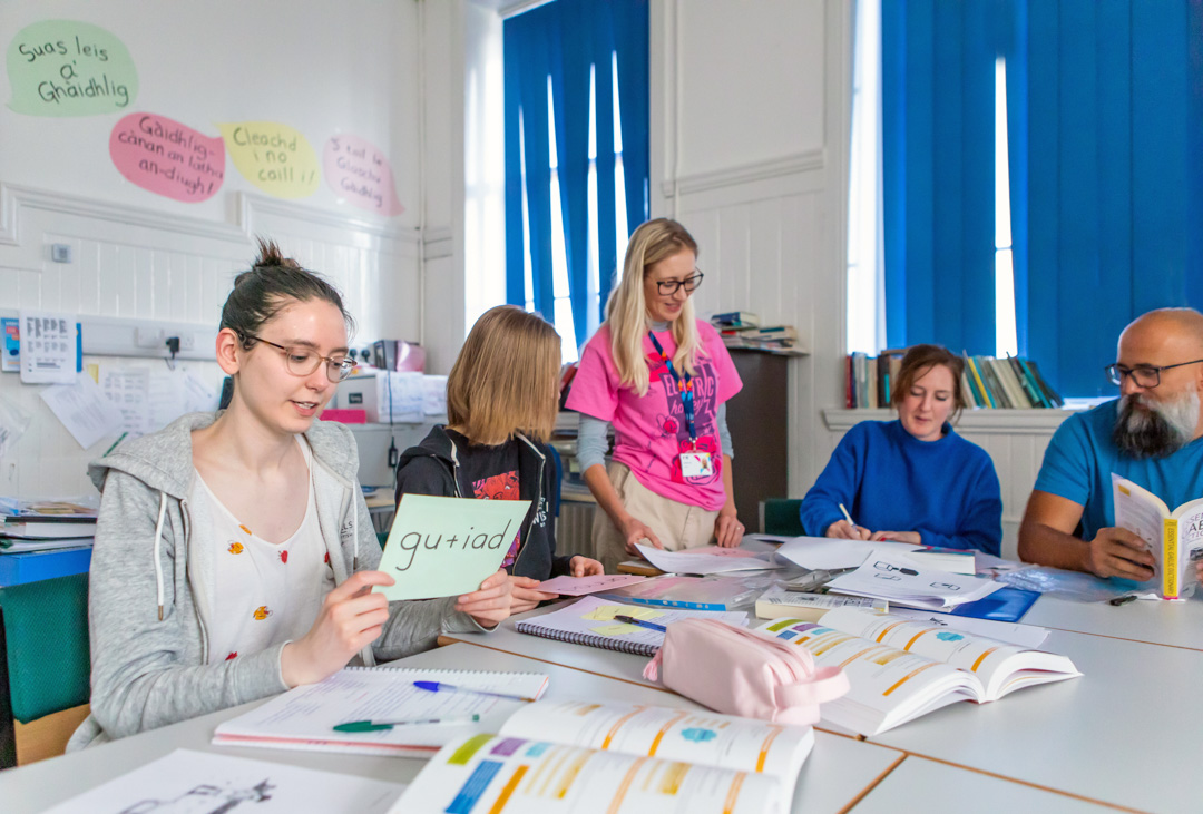 Gaelic lecturer supporting students in a language learning activity, with students holding vocabulary cards and engaging in conversation.