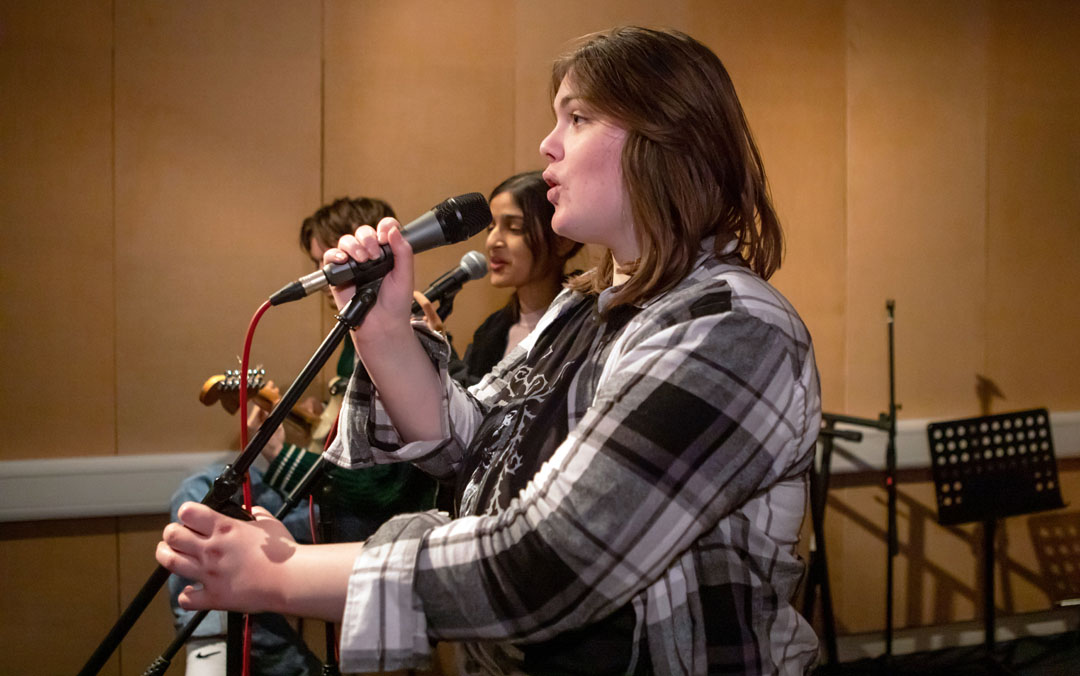 A close-up of a young singer holding a microphone and performing with a student band in a rehearsal room.