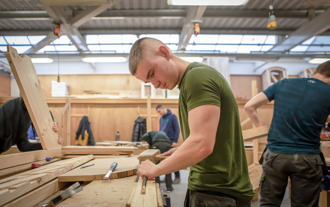 A joinery student focused on chiseling wood, using a mallet at a workbench during practical training in a woodworking class.