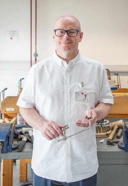 Lecturer David Webster stands in a jewellery workshop at Glasgow Kelvin College, smiling and holding a jeweller’s saw, with tools and benches in the background.