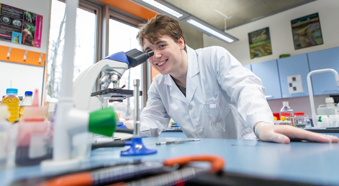 Student using a microscope in a science classroom filled with lab equipment and colourful bottles.