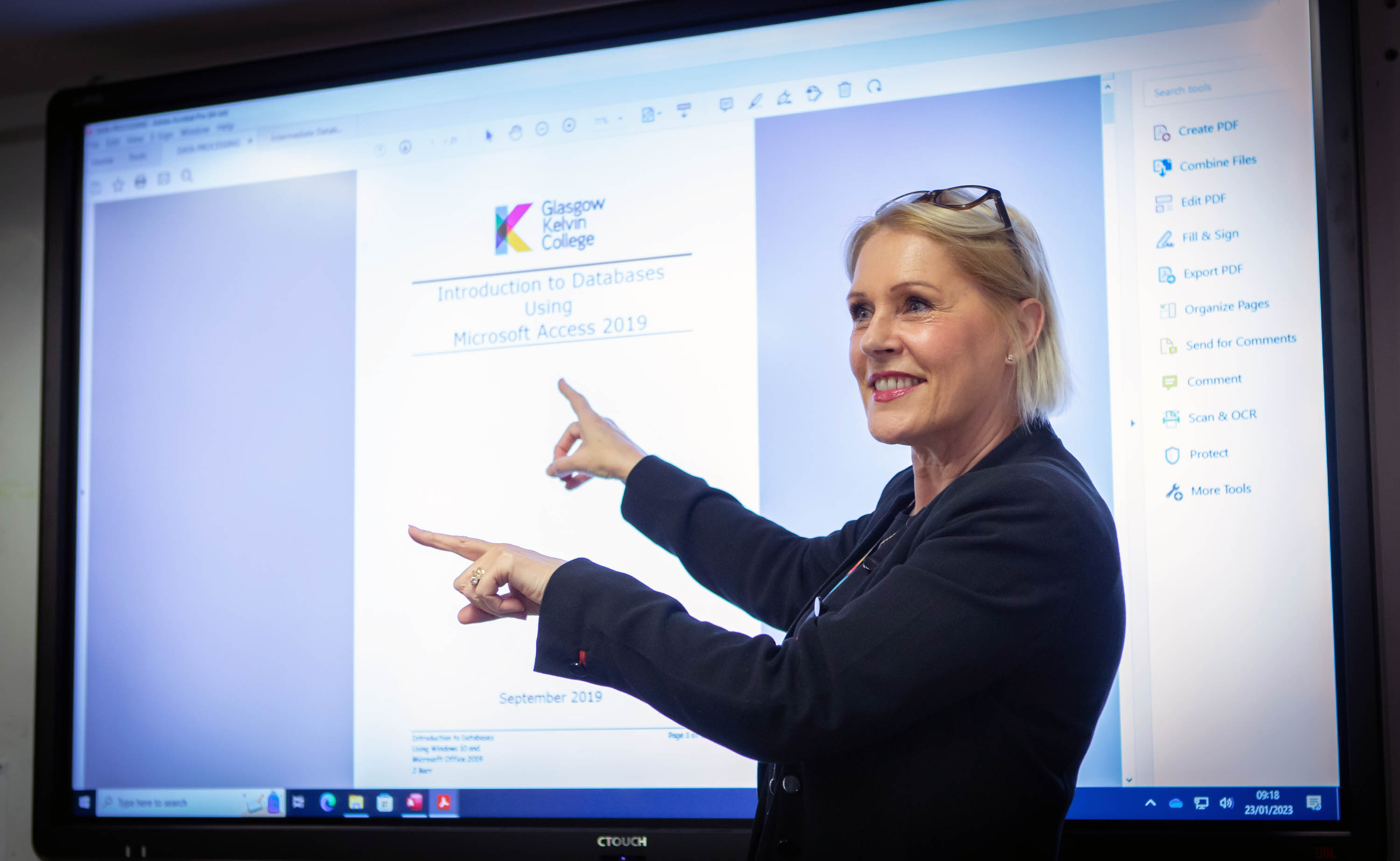 Lecturer in a black blazer presenting a lesson on Microsoft Access, pointing at a large interactive screen in a computer lab.