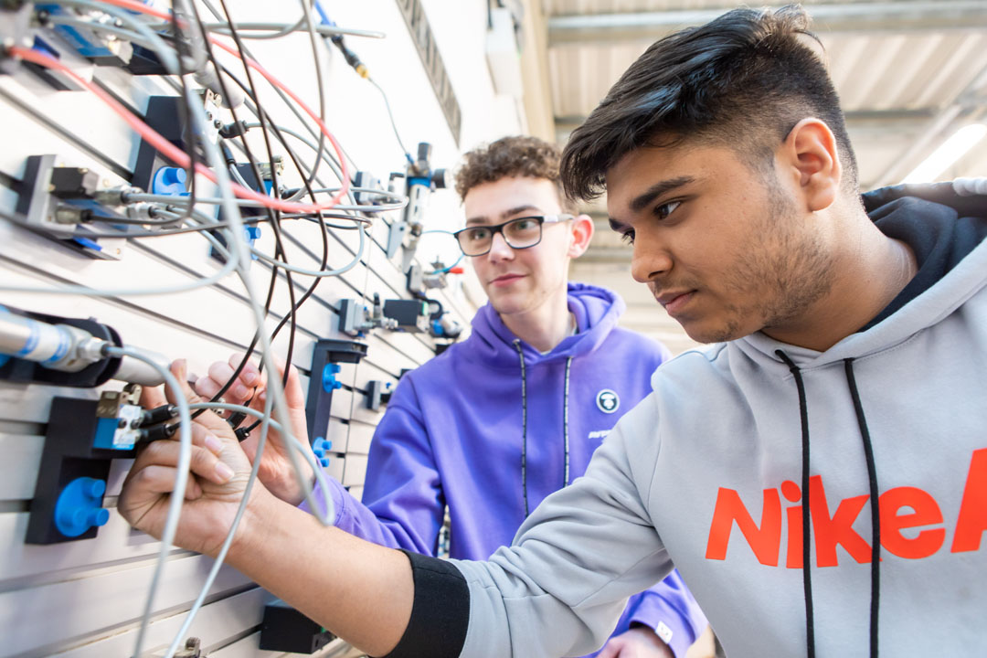 Two students working together on a pneumatics system, connecting wires and tubes to an industrial training panel.