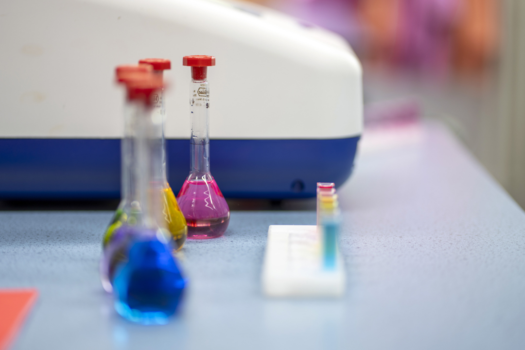 Three glass flasks filled with bright blue, yellow, and pink liquids sit on a lab bench, with a row of colourful test tubes and a science machine in the background—ready for a splash of science fun and discovery.