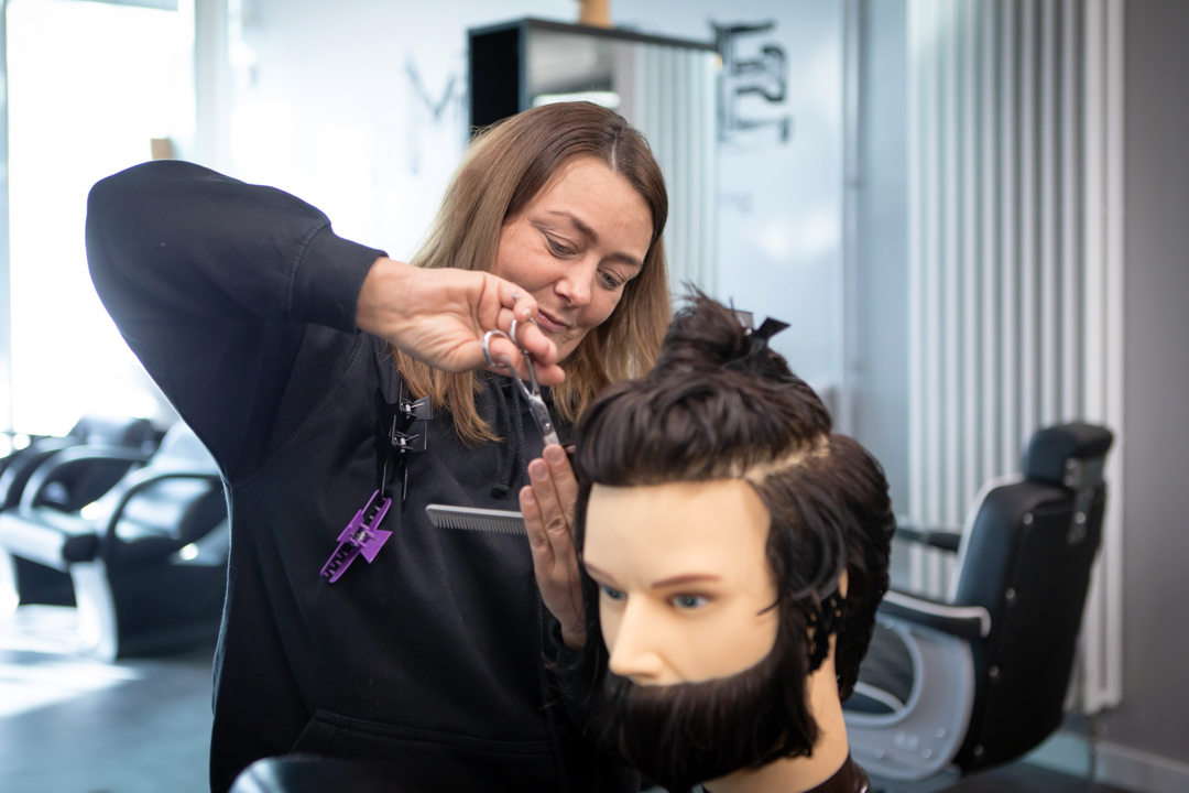 A female barbering student in a black hoodie carefully cutting the hair on a mannequin head inside a training facility.