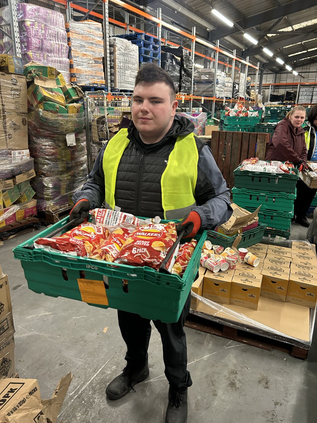 Young man wearing a high-visibility vest holding a crate of packaged crisps while working in a warehouse environment.