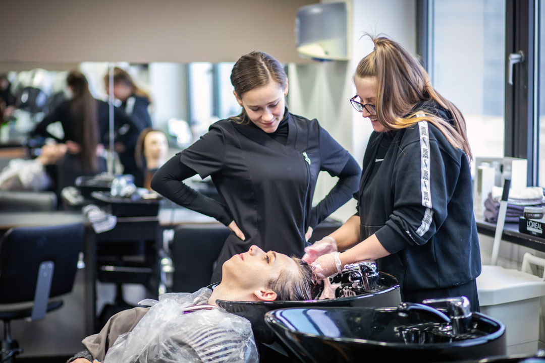 Two hairstyling students washing a client's hair at a salon sink.