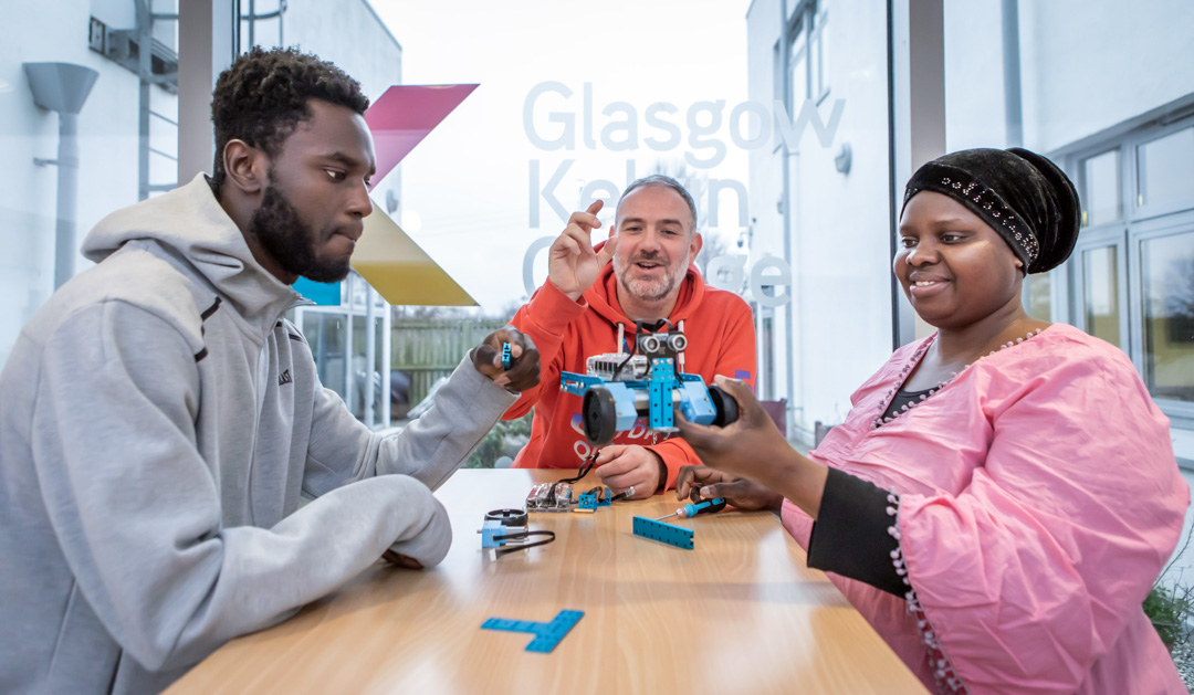 Lecturer in an orange hoodie helping two students troubleshoot and assemble a blue robotic vehicle in a classroom.