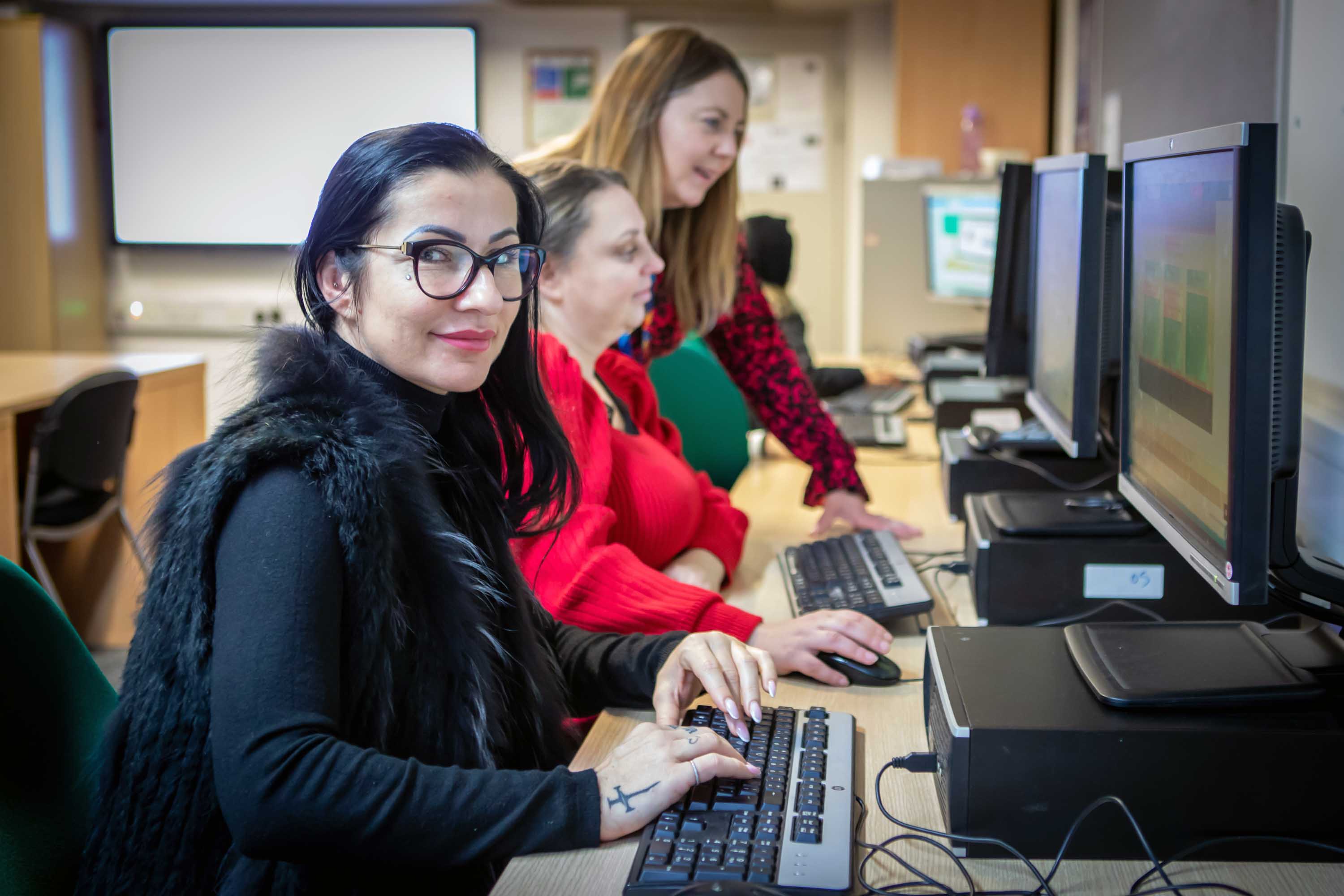 Student in a black vest working at a computer while a lecturer provides guidance and feedback in a classroom setting.