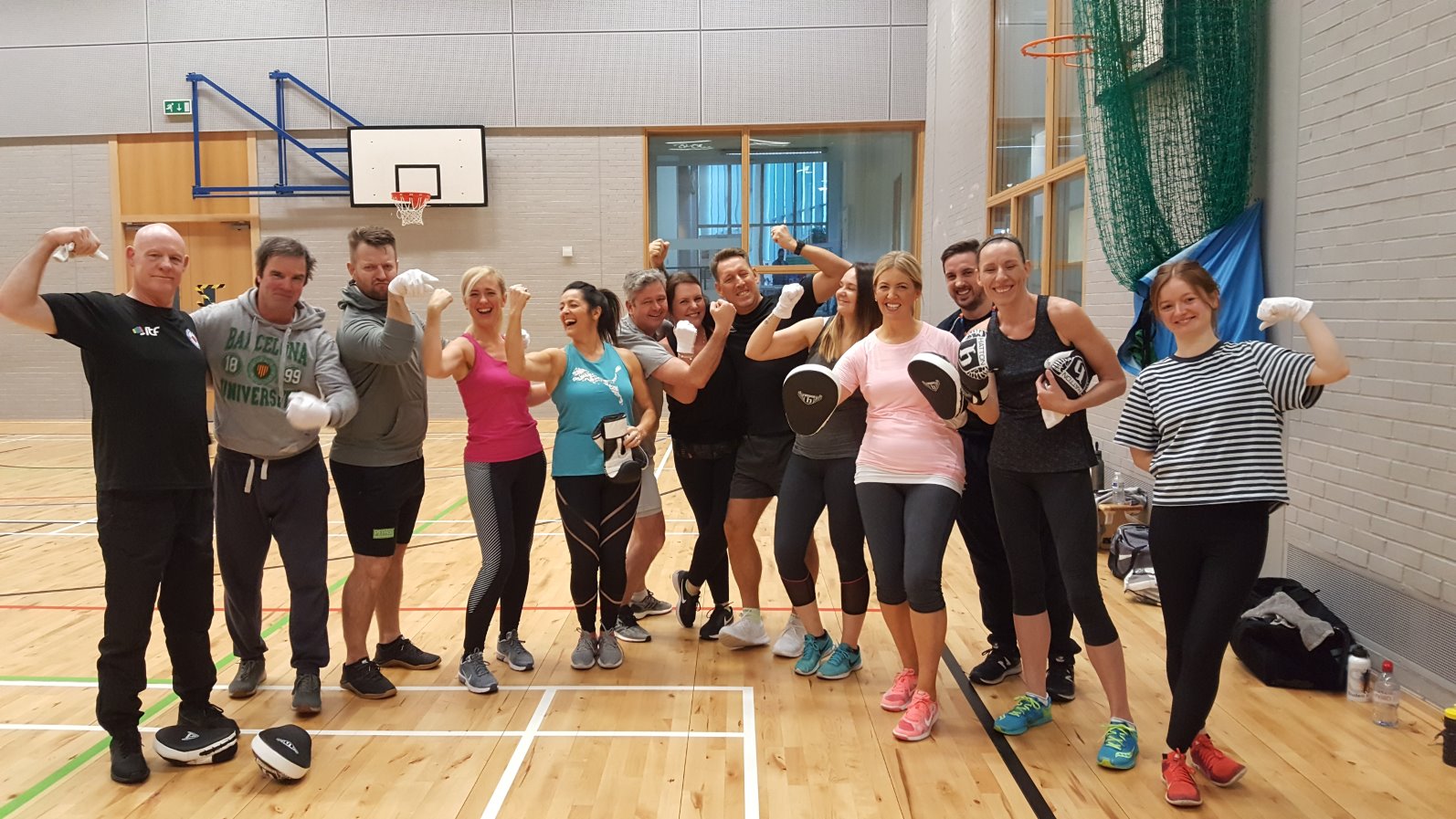 Group of Glasgow Kelvin College staff and students smiling and flexing after a fitness session in the gym hall, wearing boxing gloves and pads.
