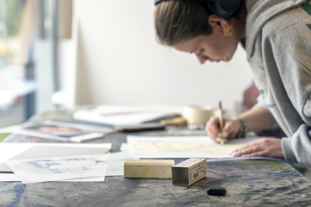 Person leaning over a table, drawing on paper in an art studio, with various sketches and art materials spread out