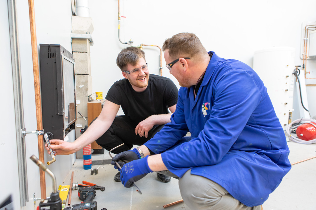 A plumbing lecturer and student smiling and collaborating closely while working together to install copper piping in a practical training session.