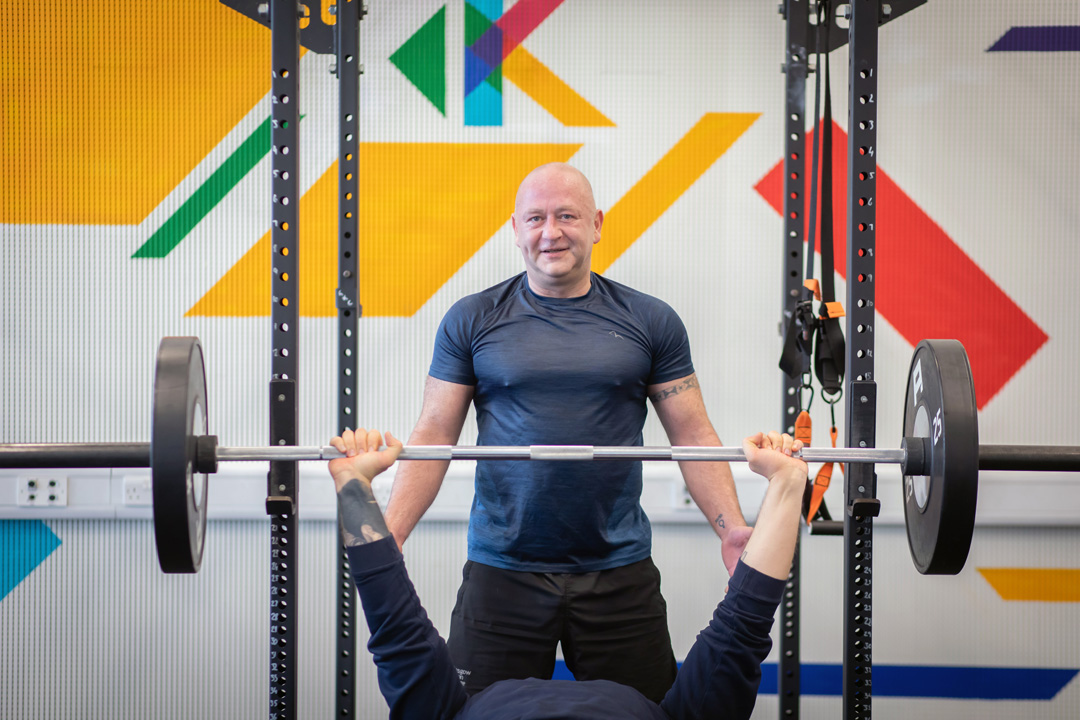 Student performing a bench press while another student spots them in a colourful training area.
