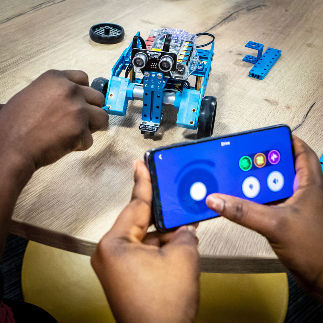 Student using a smartphone to control a small blue robotic vehicle, testing its movements on a table in a computing lab.