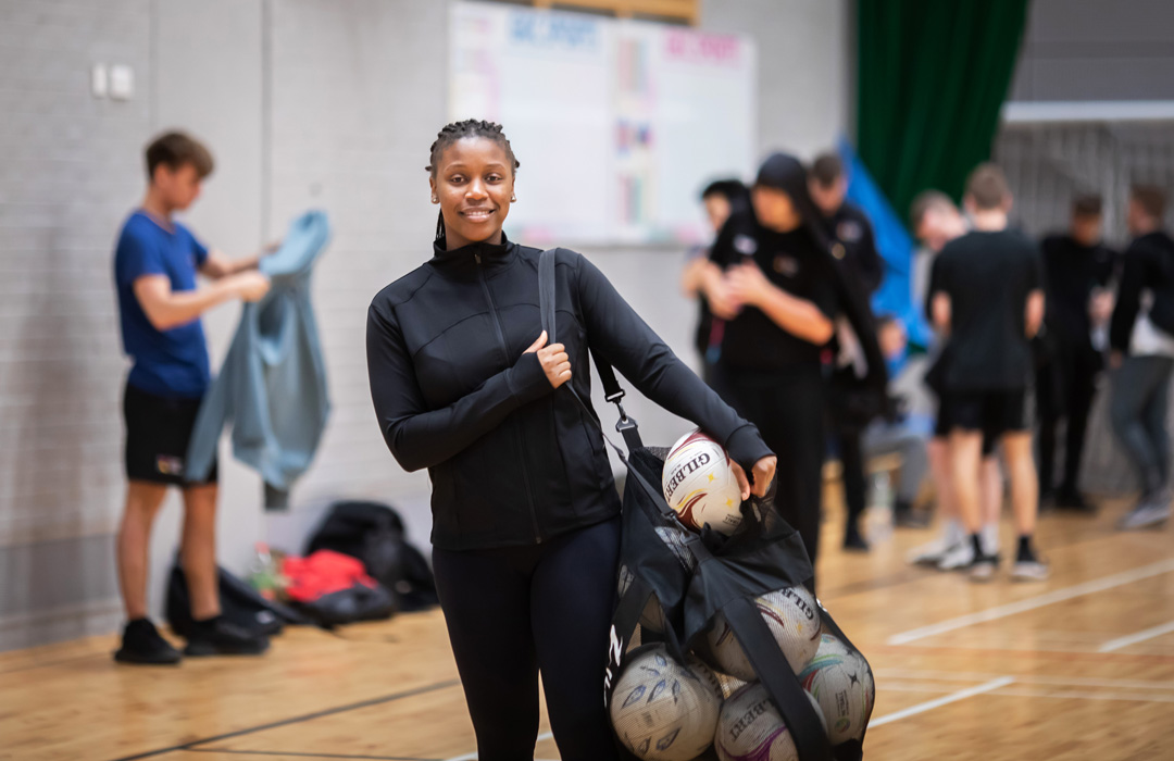 A student holding a mesh bag filled with netballs, smiling at the camera in a sports hall.