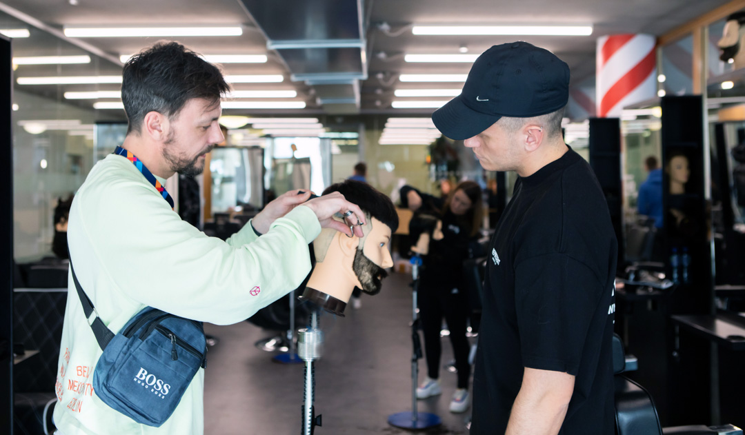 A barbering instructor demonstrating a haircut on a mannequin head while a student in a black cap observes.