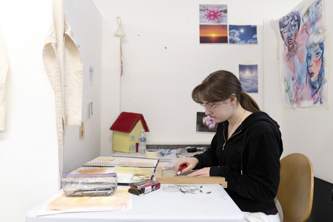 Person seated at a desk in an art studio, carving a design into a printmaking block, with sketchbooks and artwork on the wall