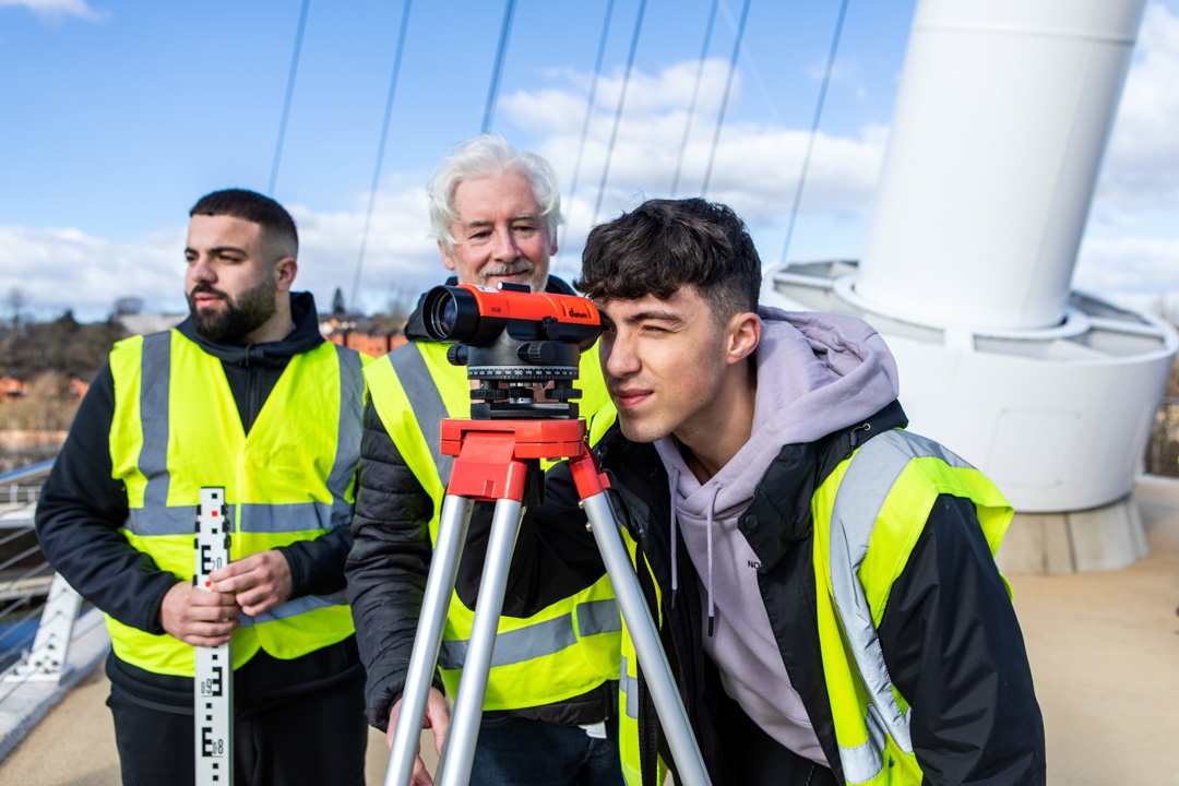A young student in a high-visibility vest looking through a surveying instrument while the lecturer and another student observe and discuss measurements on a bridge