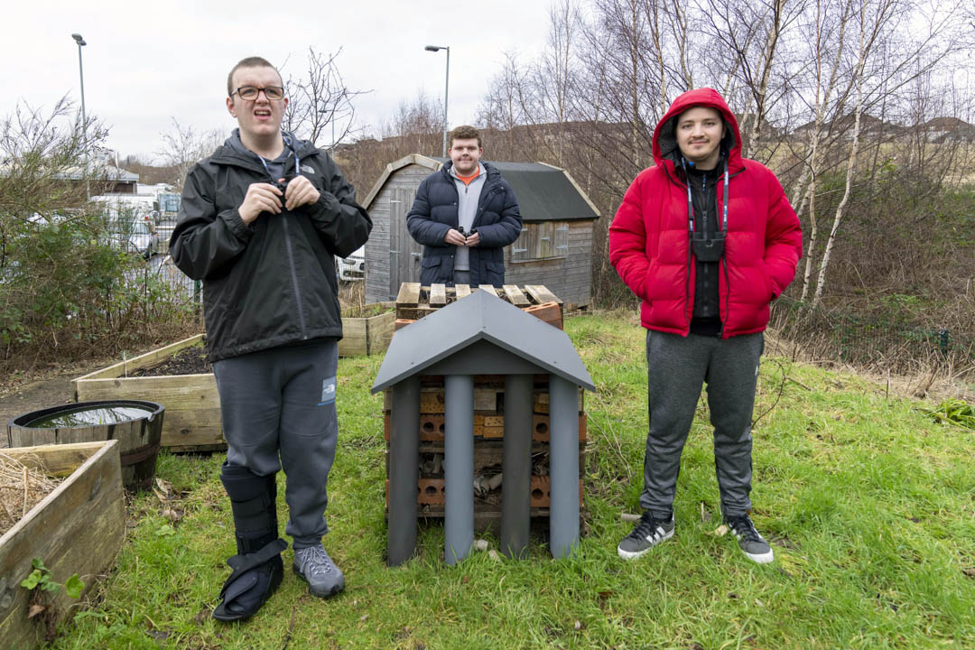 Three young men standing outside beside a handmade bug hotel, surrounded by greenery and raised garden beds.