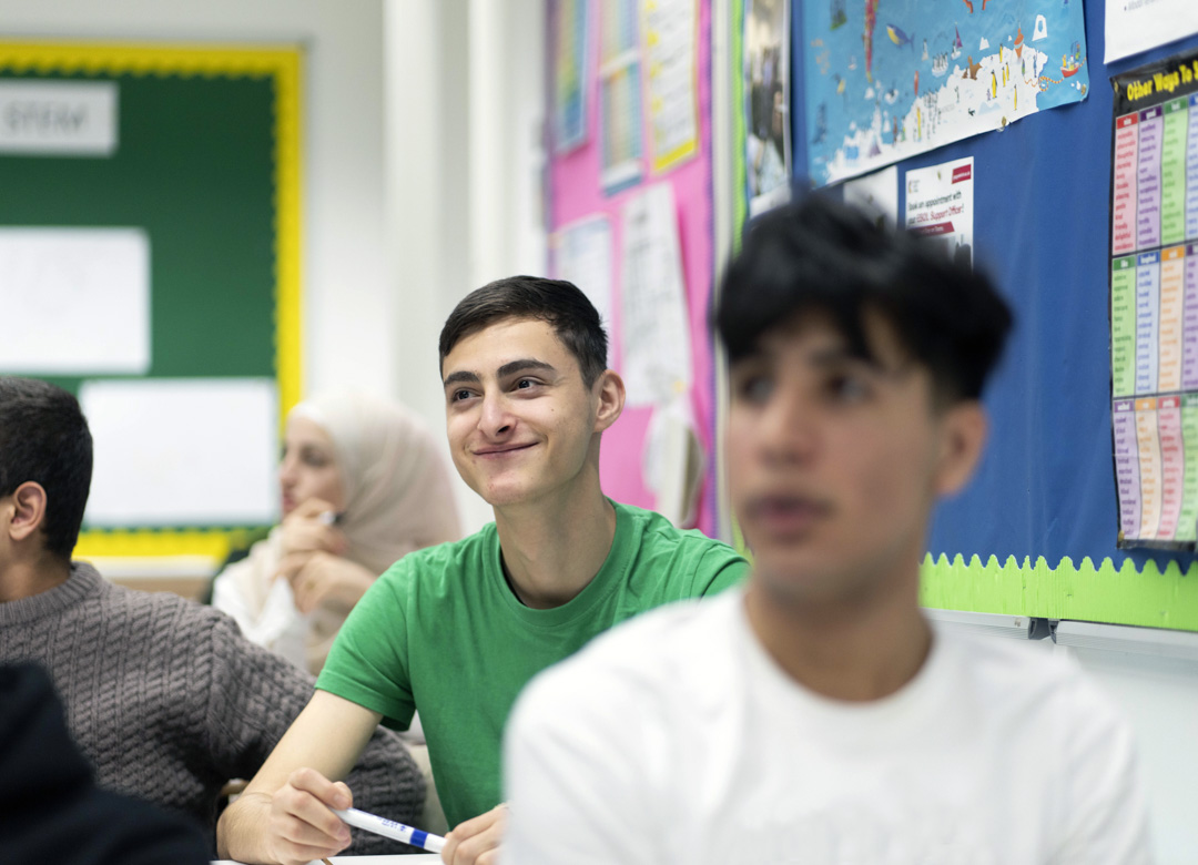 A young male student in a green t-shirt sits in a classroom, smiling while holding a whiteboard marker. Other students are visible in the background, including a student in a white hijab.