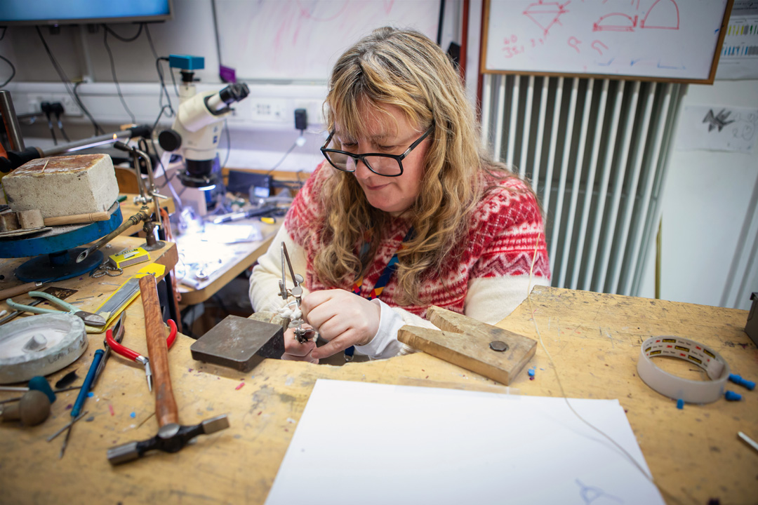 A female jewellery lecturer with long blonde hair and glasses, wearing a red and white sweater, working at a jewellery-making bench in a workshop filled with tools and equipment.