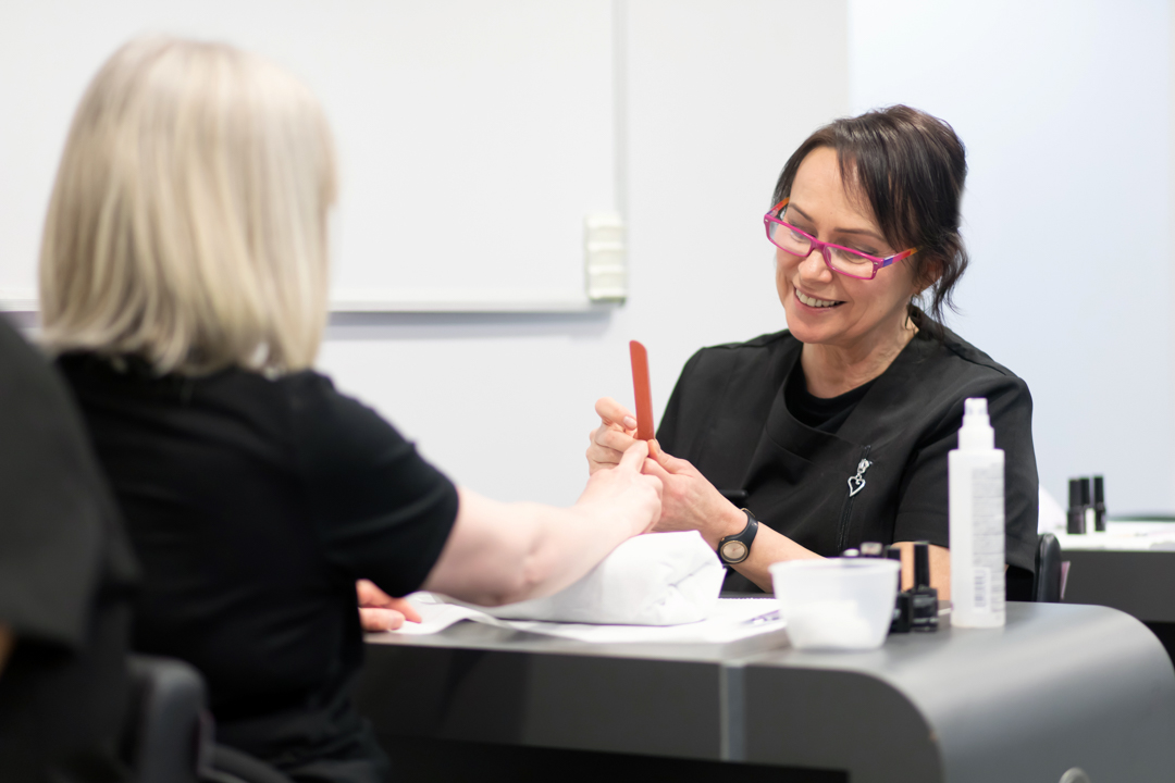 A beauty therapy student provides a manicure to a client, shaping nails with a file while smiling in a professional training setting.