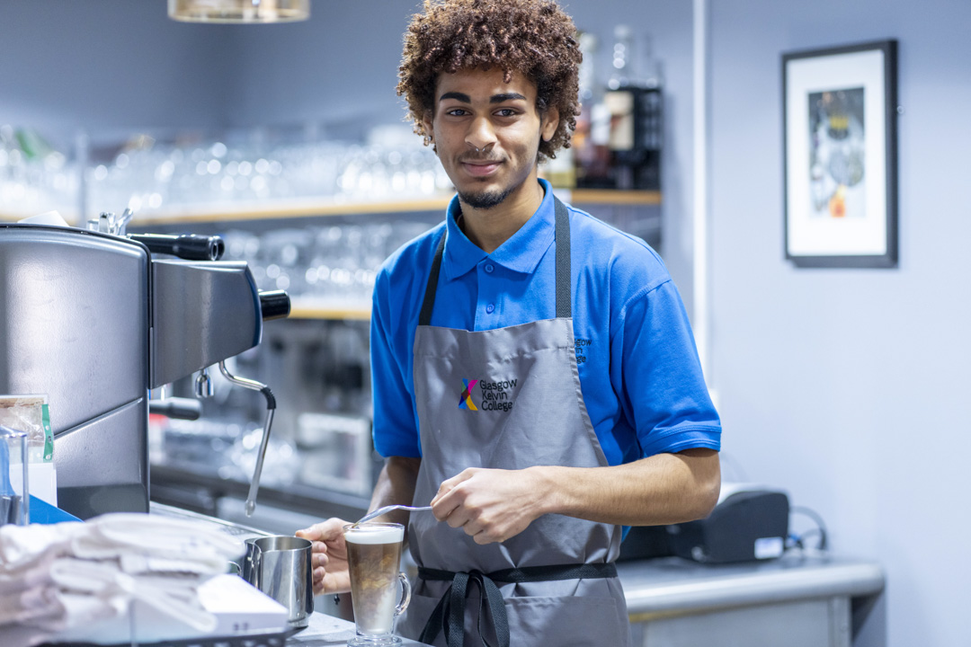 A young male barista in a blue uniform and grey apron, smiling while preparing a latte at a coffee machine in a café setting.