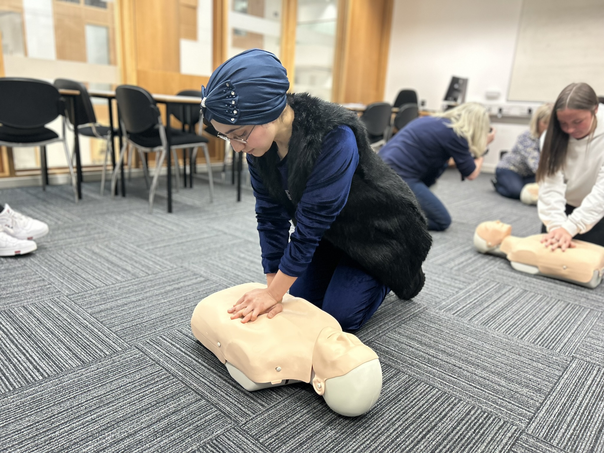 A woman wearing a blue headscarf and glasses practices CPR on a mannequin during a first aid training session in a classroom setting. Other participants are visible in the background.