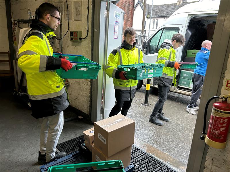 Three young men in high-visibility jackets unloading green crates from a delivery van at a warehouse entrance.