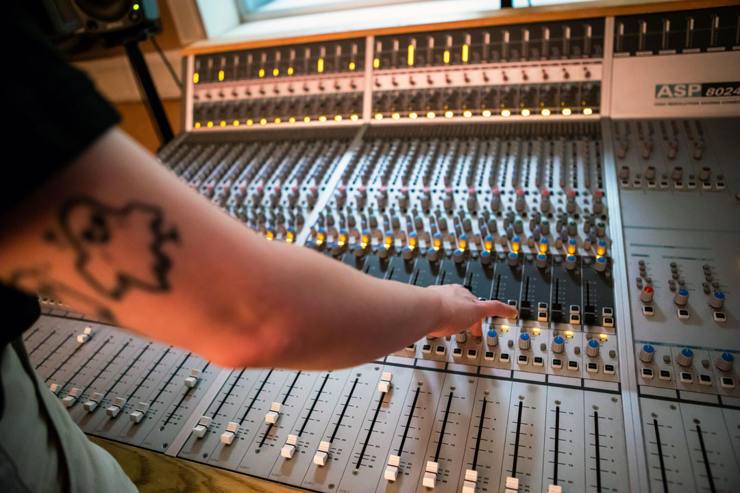 A close-up of a student's arm with a ghost tattoo reaching for controls on a large mixing desk, with glowing indicator lights.