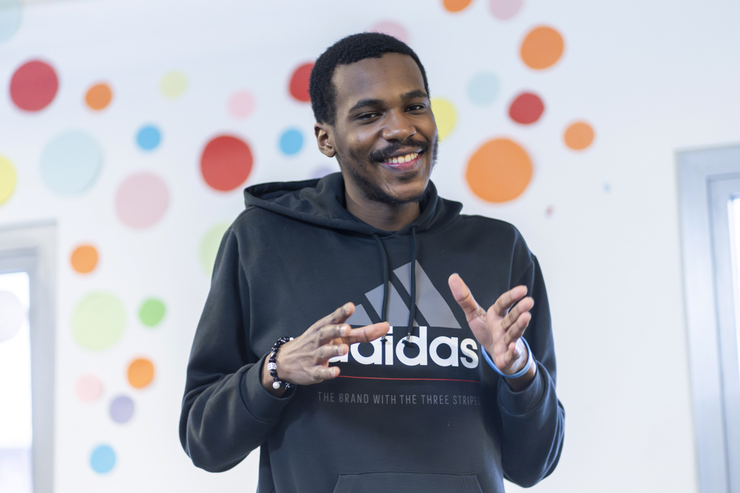 Young man smiling while making expressive hand gestures in front of a colorful polka-dot wall.