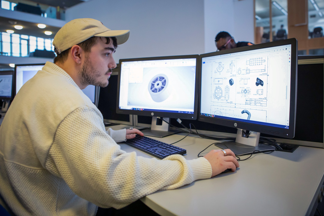 A student wearing a white cap working on a dual-monitor CAD setup, designing a mechanical component on one screen and a technical drawing on the other.