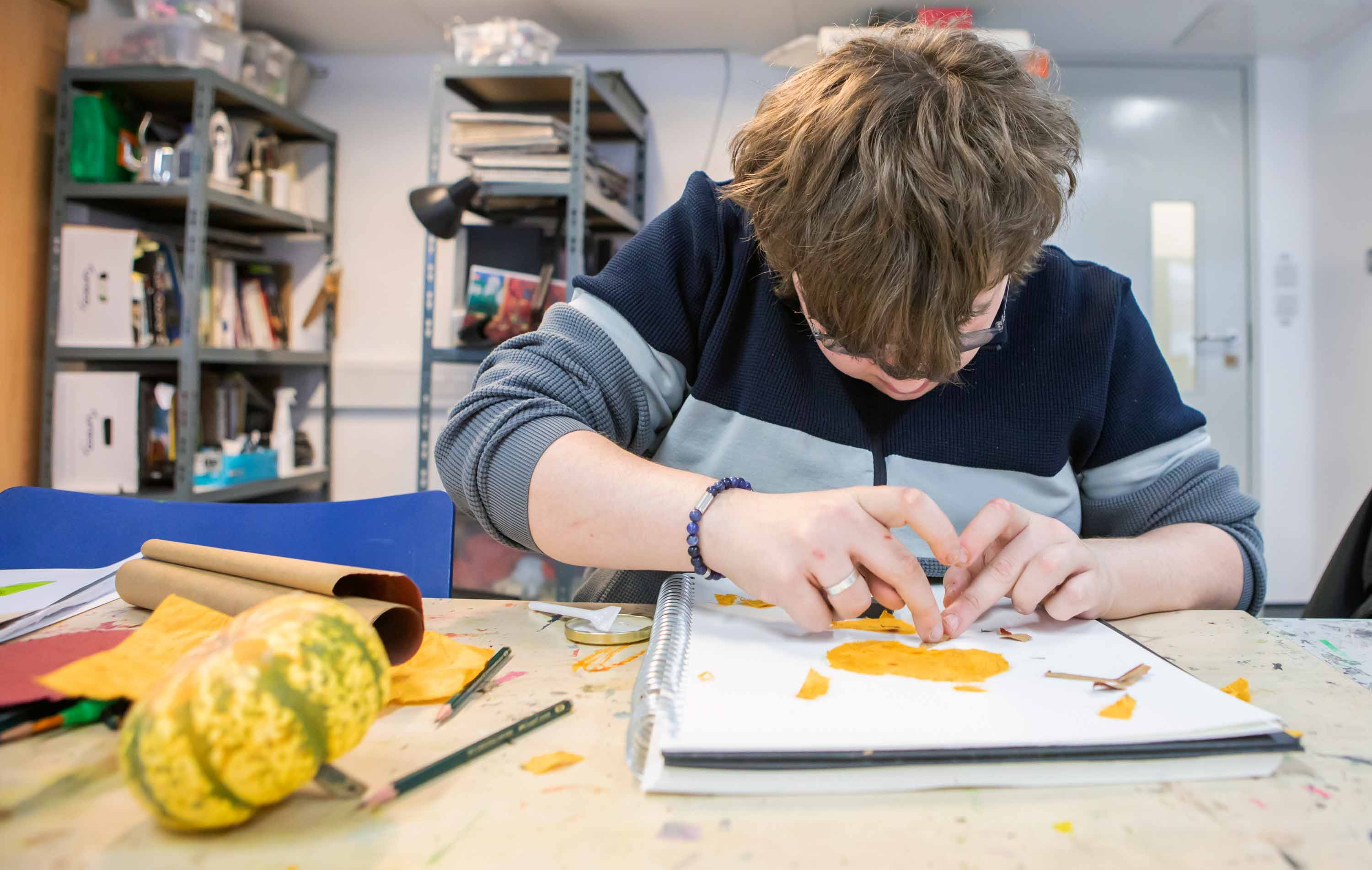 Student working on an art project, using orange paper to create a design in a sketchbook, surrounded by art supplies.