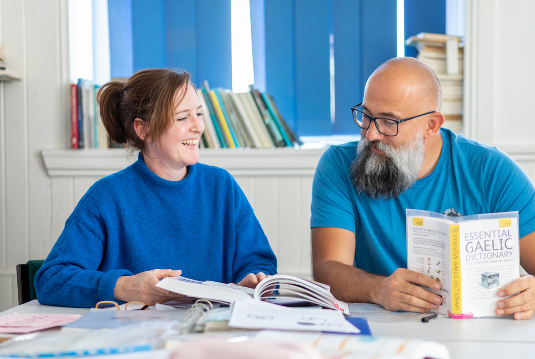 Two students in a Gaelic language class laughing while studying, with a Gaelic-English dictionary open on the desk.