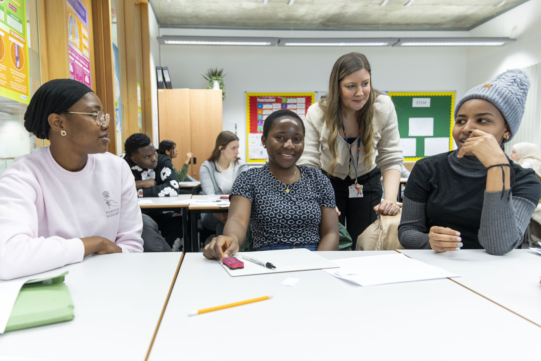 A classroom scene where three female students are smiling and engaging in discussion. A teacher in a beige cardigan leans over the table to interact with them, while other students work in the background.